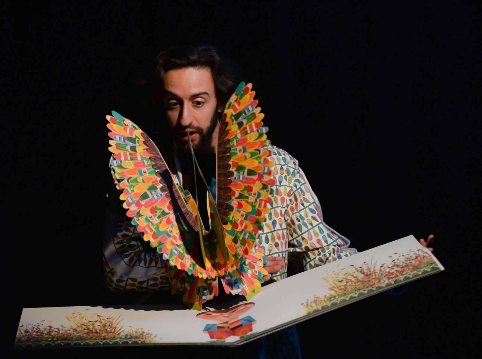 Artista em cena com um livro ilustrado e um pássaro de muitas cores em papel