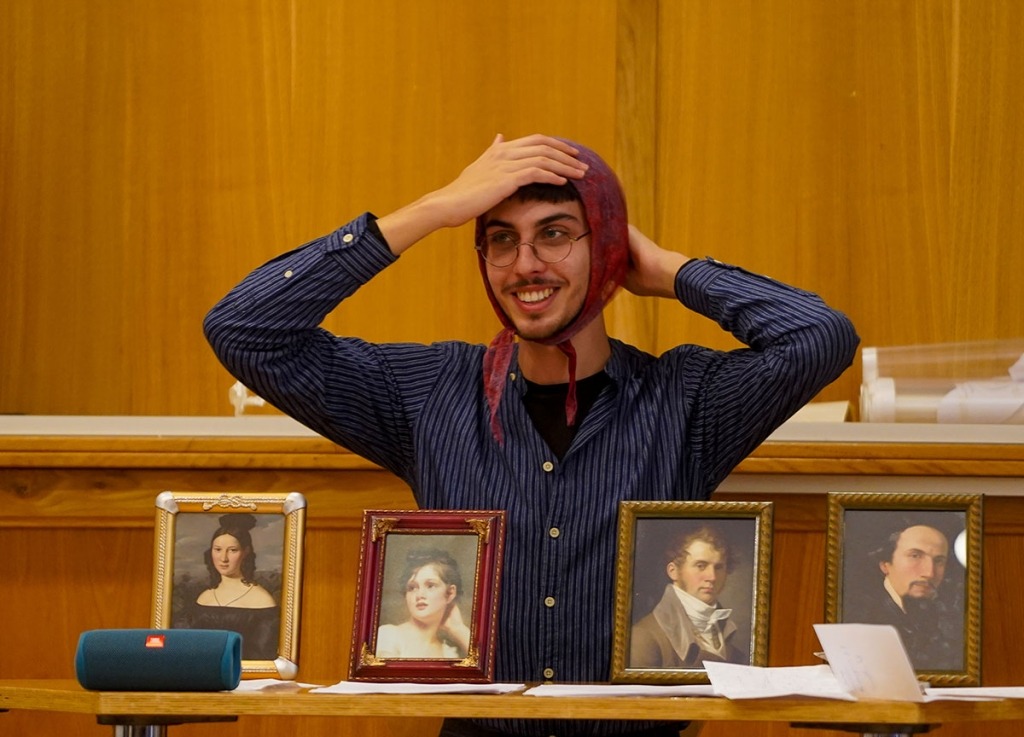 um homem ainda jovem com um lenço na cabeça imita os retratos nas molduras que tem à frente