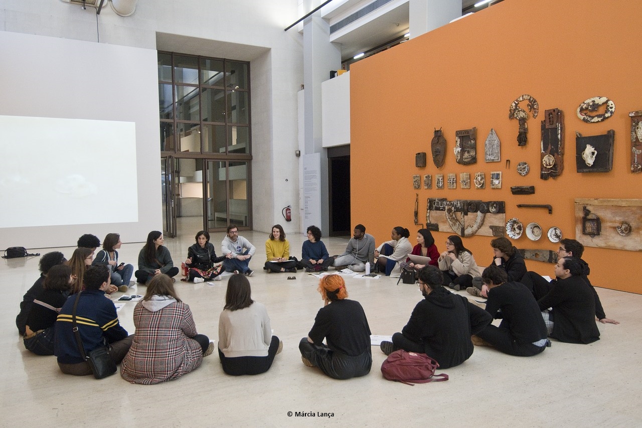 Várias pessoas sentadas em forma de roda, no chão de um museu