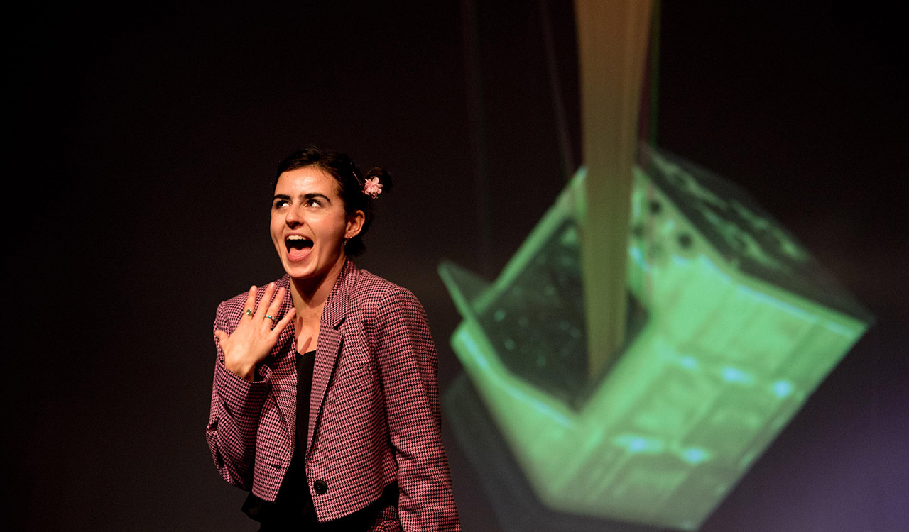 Mulher sorri de forma exagerada com um mão junto ao peito; atrás de si uma projeção de um forno de cozinha