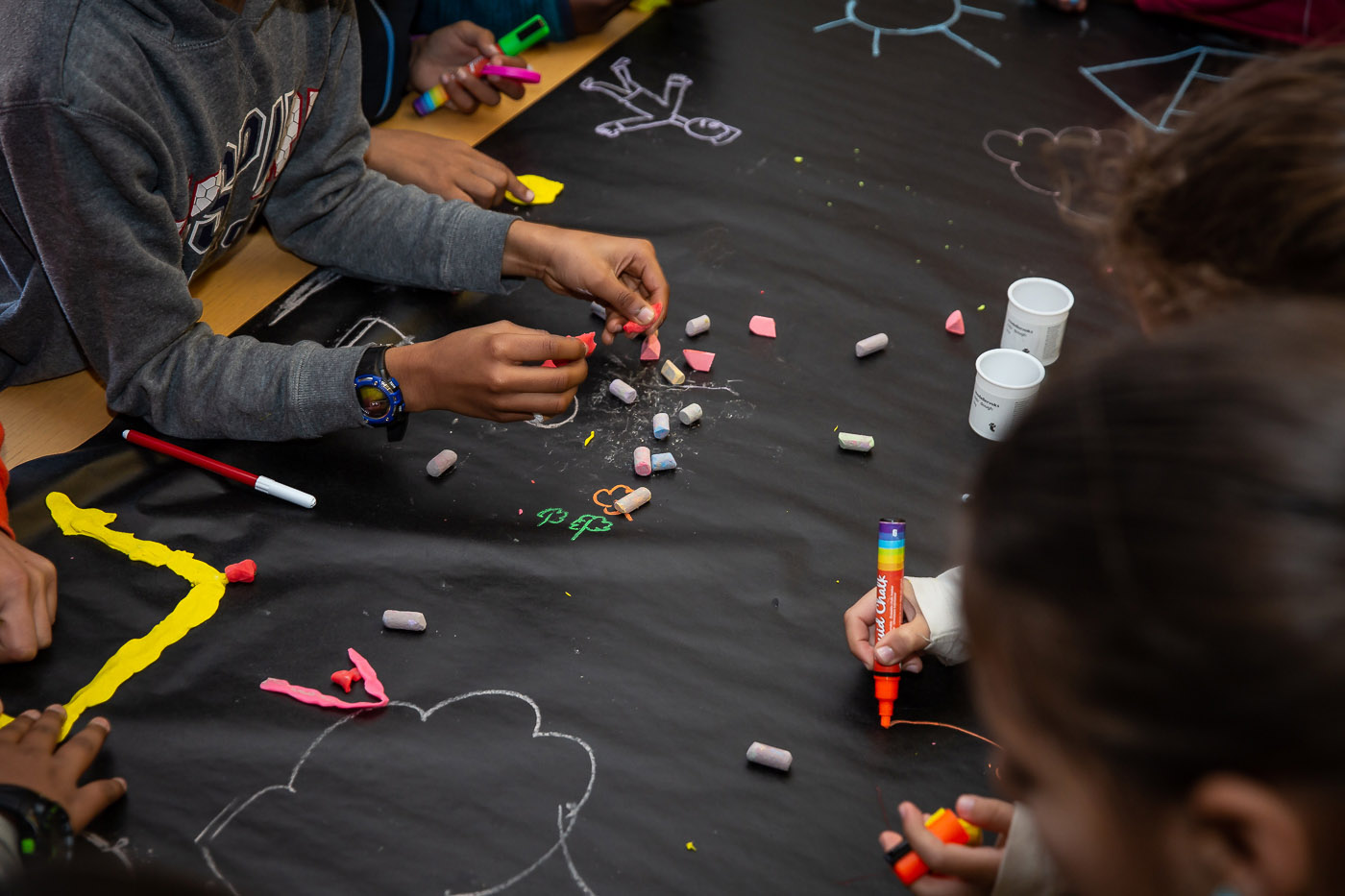 Mãos pegam em paus de giz coloridos numa mesa forrada com cartolina preta